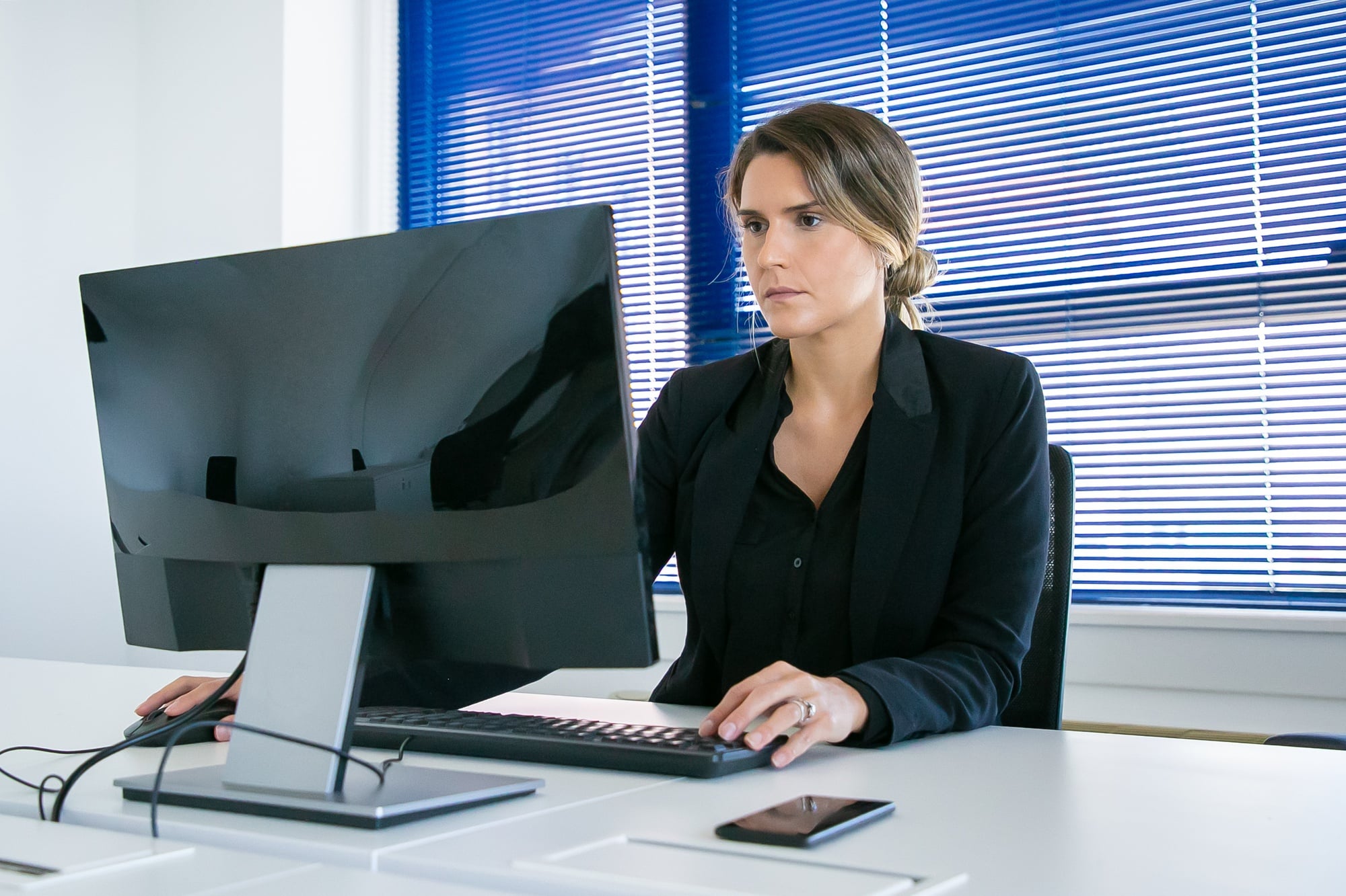 A woman in a black blazer sits at a desk working on a desktop computer with secure cloud connectivity in a bright office with blue blinds covering the windows in Minnesota. A smartphone and documents are on the desk.