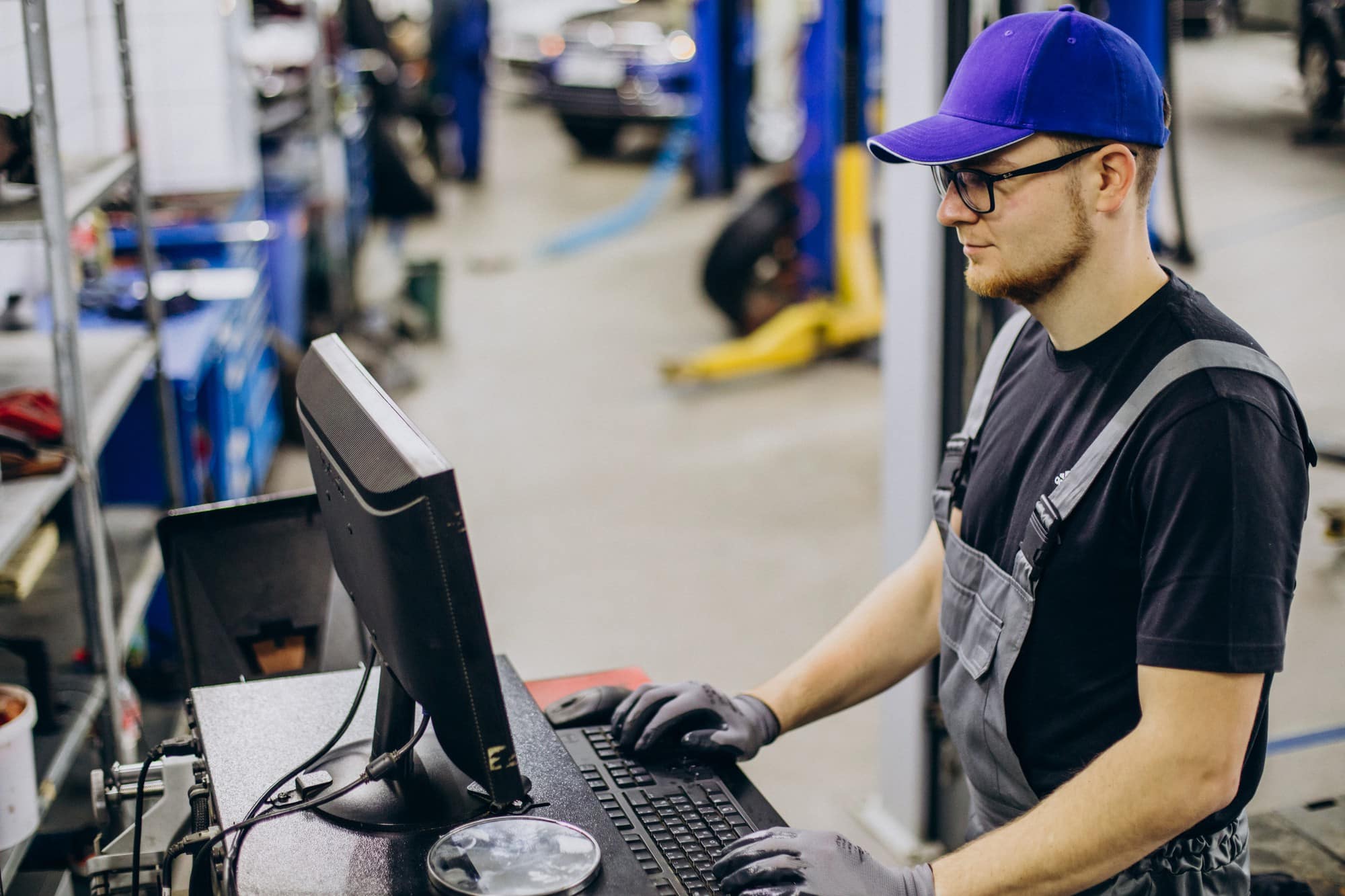 A mechanic wearing a blue cap, glasses, gloves, and overalls in a manufacturing firm in Minnesota, types on a computer keyboard with secure cloud connectivity.