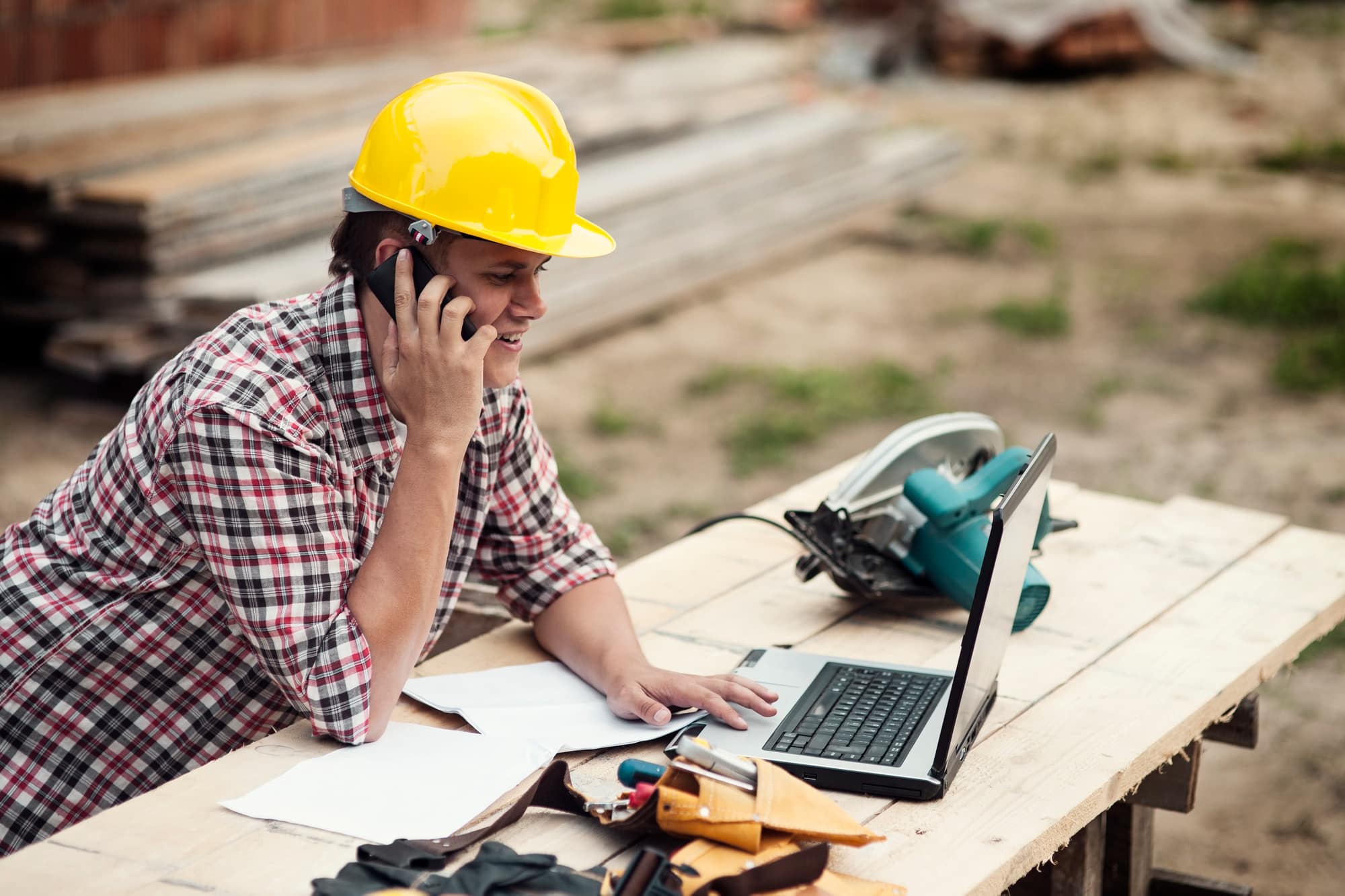 A construction worker wearing a yellow hard hat and plaid shirt talks on the phone while using a laptop at a wooden table outdoors, with papers, tools, and a circular saw nearby at a Minnesota job site.