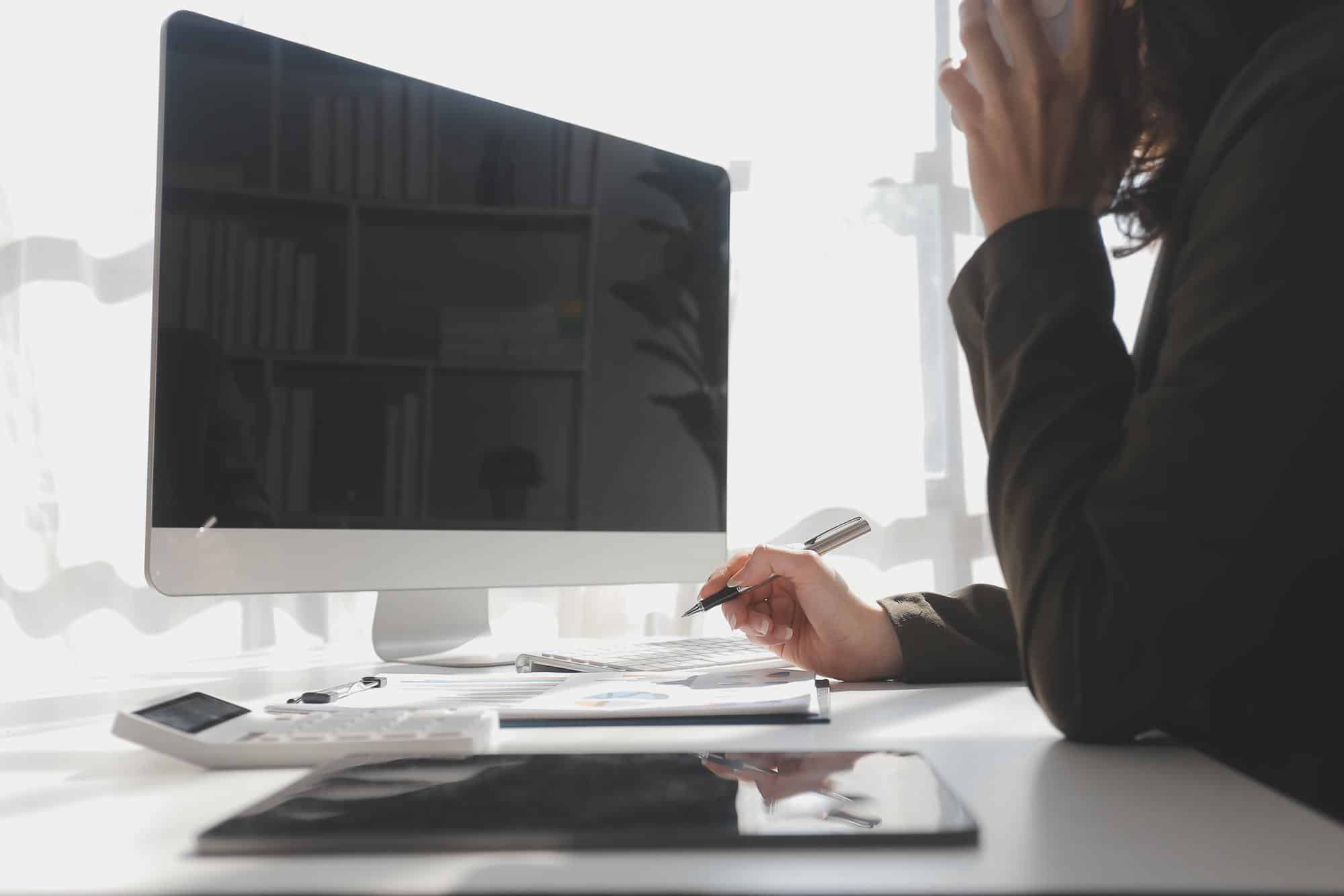 A person sits at a desk in front of a computer monitor providing managed IT support for businesses in Minnesota, holding a pen and speaking on the phone. Office supplies, papers, a calculator, and a tablet are on the desk. Sunlight filters through a window.