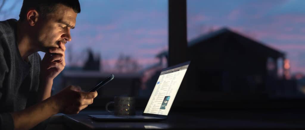 A man sits at a desk in a dimly lit room, looking thoughtfully at his phone while browsing for solutions for his IT problems and unreliable tech support. A laptop displaying a website is open in front of him. A mug is on the table and a window shows a sunset outside.